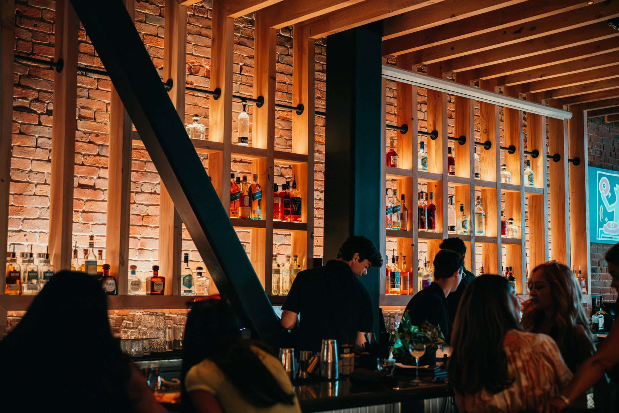 Dimly lit bar with illuminated, backlit liquor bottles displayed on wall shelving and silhouetted patrons at the counter.