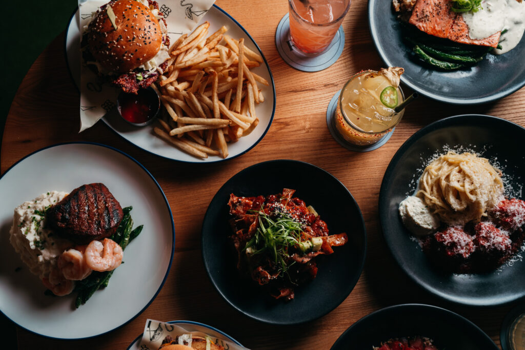  Overhead view of restaurant dishes: burger and fries, seared meats, fish with vegetables, and desserts with cocktails on a wooden table.