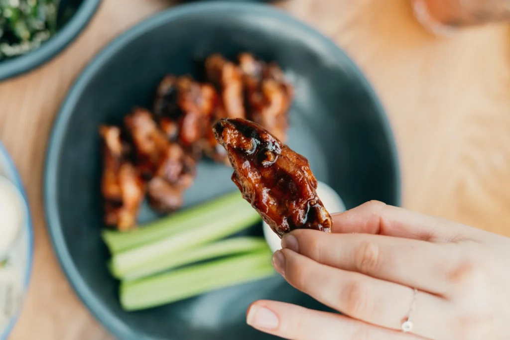 Hand holding a glossy glazed chicken wing over a bowl of cooked wings and green onions.